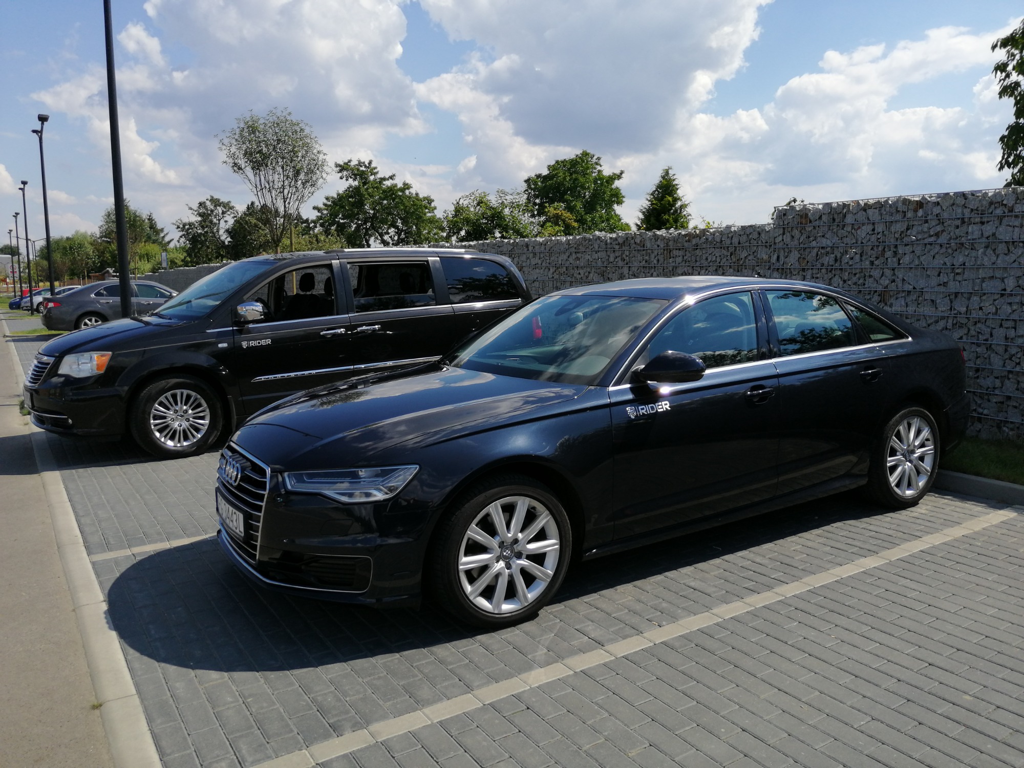 Dwa czarne samochody osobowe, minivan Chrysler Town & Country i Audi A6, zaparkowane na parkingu z kostki brukowej, z logo firmy RIDER na karoserii, w tle mur z gabionów i błękitne niebo z obłokami.