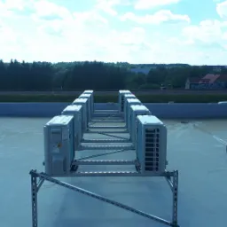 Rząd zewnętrznych jednostek klimatyzacyjnych na dachu budynku z widokiem na linię kolejową i zieleń w tle.