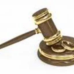 Drewniany młotek sędziowski z pozłacanymi elementami spoczywa na białym tle, obok leżą dwie złote obrączki ślubne.