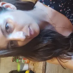 Portret kobiety z ciemnymi włosami i delikatnym łańcuszkiem na szyi, ubrana w kwiecistą bluzkę, z widocznymi butelkami i pojemnikami w tle.