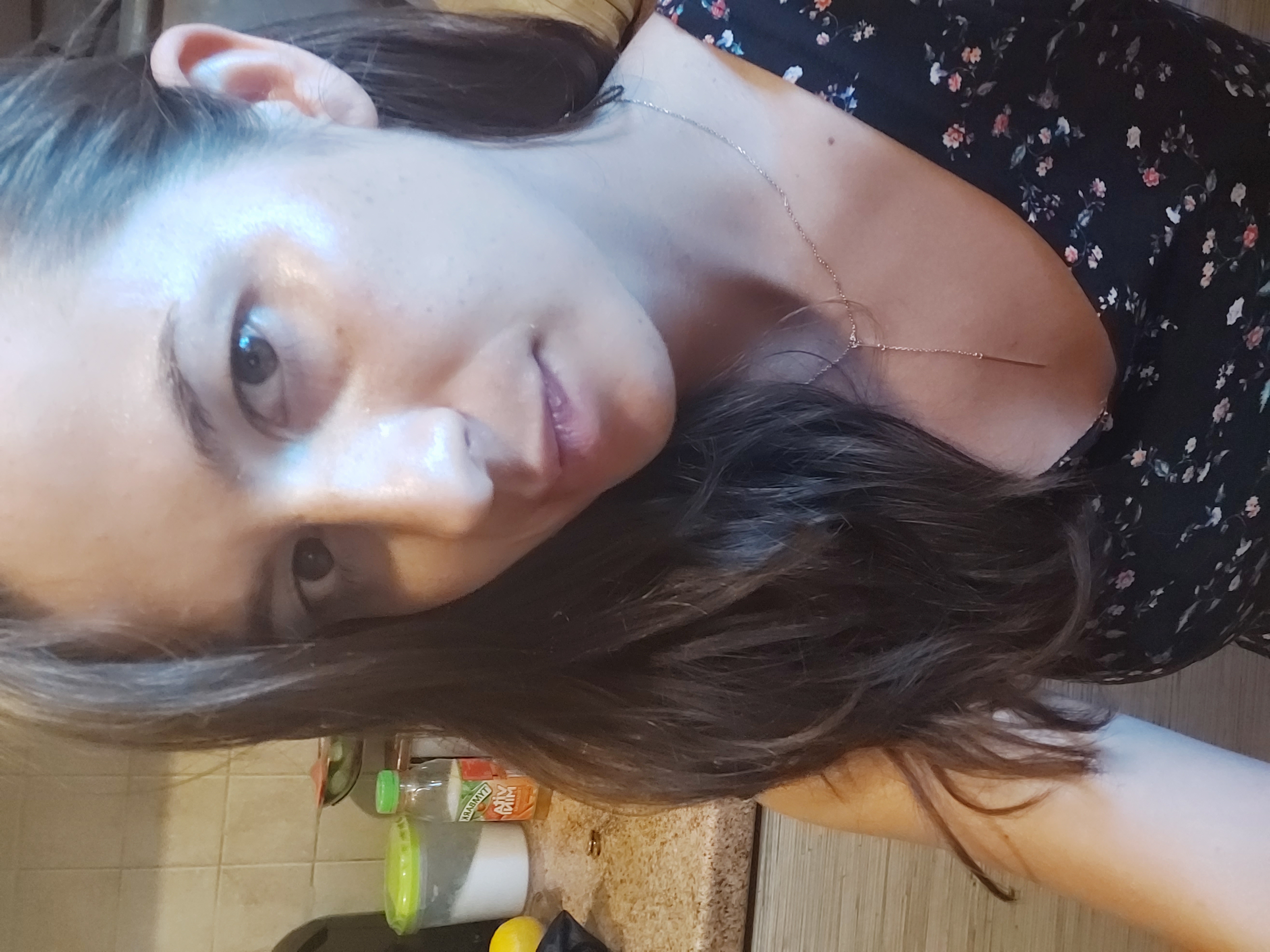 Portret kobiety z ciemnymi włosami i delikatnym łańcuszkiem na szyi, ubrana w kwiecistą bluzkę, z widocznymi butelkami i pojemnikami w tle.