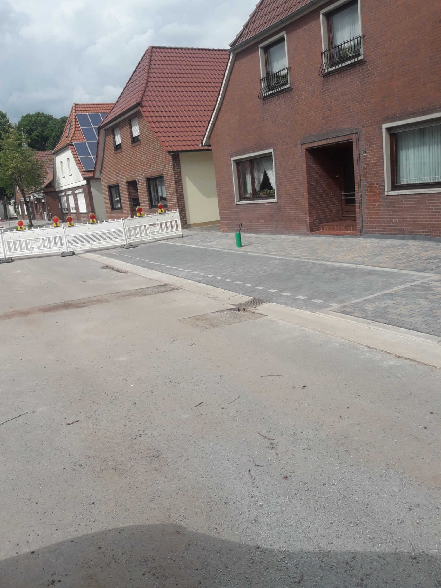 Ulica po remoncie z nową nawierzchnią z kostki brukowej w odcieniach szarości, zabezpieczona barierkami z tworzywa sztucznego, w tle budynki mieszkalne z czerwonej cegły i dachówką.