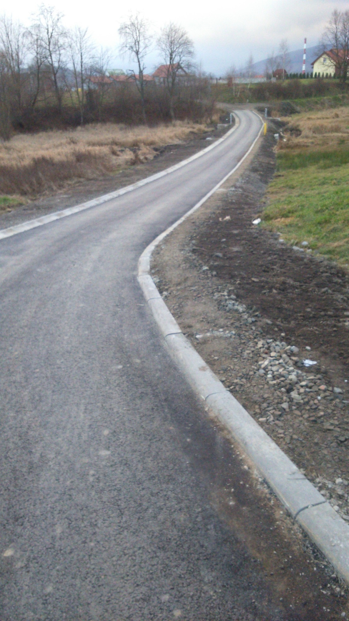 Nowo wybudowana asfaltowa droga z betonowymi krawężnikami na skraju pola w pochmurny dzień, w tle widoczne drzewa i budynki.