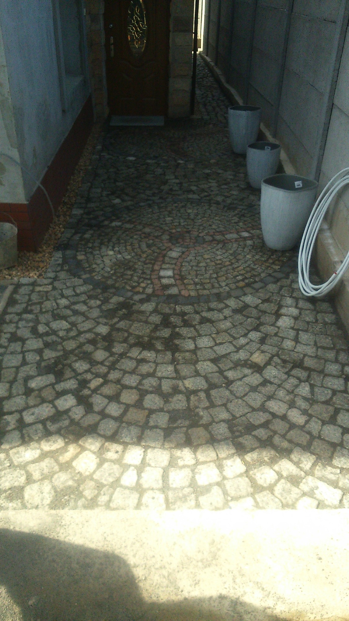 Ścieżka z kostki granitowej ułożona w dekoracyjny wzór wachlarza z czerwonym akcentem, prowadząca do drzwi wejściowych; po bokach ustawione trzy szare donice.