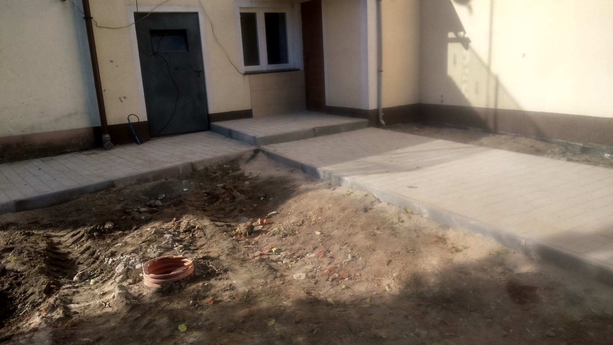 Świeżo ułożony chodnik z kostki brukowej prowadzący do drzwi budynku, widoczne niedociągnięcia w postaci ziemi i resztek budowlanych dookoła, pomarańczowa rura leżąca na ziemi.