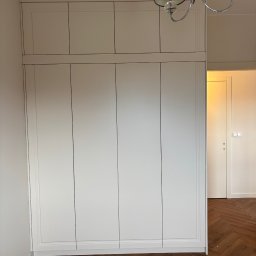 Mój.dom - Biała szafa wnękowa z górnymi szafkami, widoczna na tle parkietu ułożonego w jodełkę. W tle widoczne drzwi do innego pomieszczenia i ozdobny żyrandol.