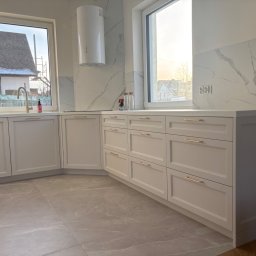 Mój.dom - Jasna kuchnia z białymi frontami i złotymi uchwytami, marmurowy backsplash, widok na zewnątrz przez okno. Nowoczesny design z elementami klasycznymi.