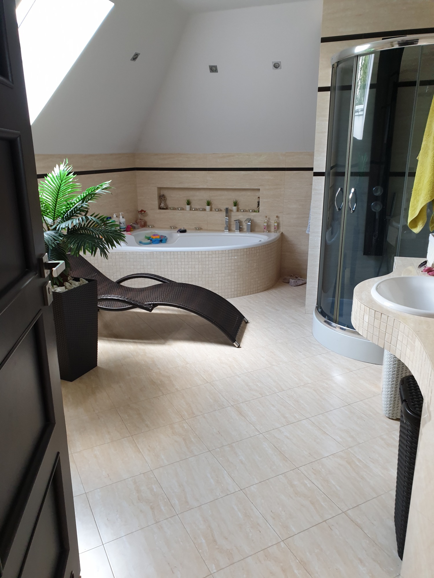 Jasna łazienka na poddaszu z beżowymi płytkami, wanną z hydromasażem, kabiną prysznicową i leżanką z rośliną doniczkową, widok na otwarte drzwi.