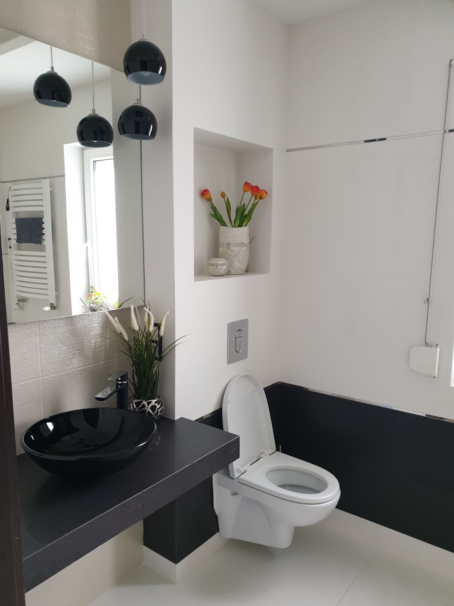 Nowoczesna łazienka z czarną umywalką na blacie, wiszącą toaletą i dekoracyjną wnęką z kwiatami, oświetlona trzema czarnymi lampami wiszącymi.