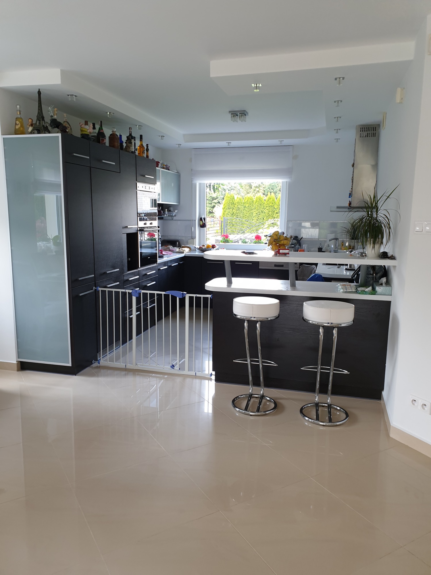 Nowoczesna, jasna kuchnia z ciemnymi szafkami, białym blatem, dwoma chromowanymi hokerami i bramką zabezpieczającą dla dzieci.