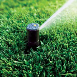 Niskie ujęcie czarnego zraszacza w trawie, wyrzucającego strumień wody pod kątem, z widocznymi detalami głowicy i zielonym tłem trawnika.