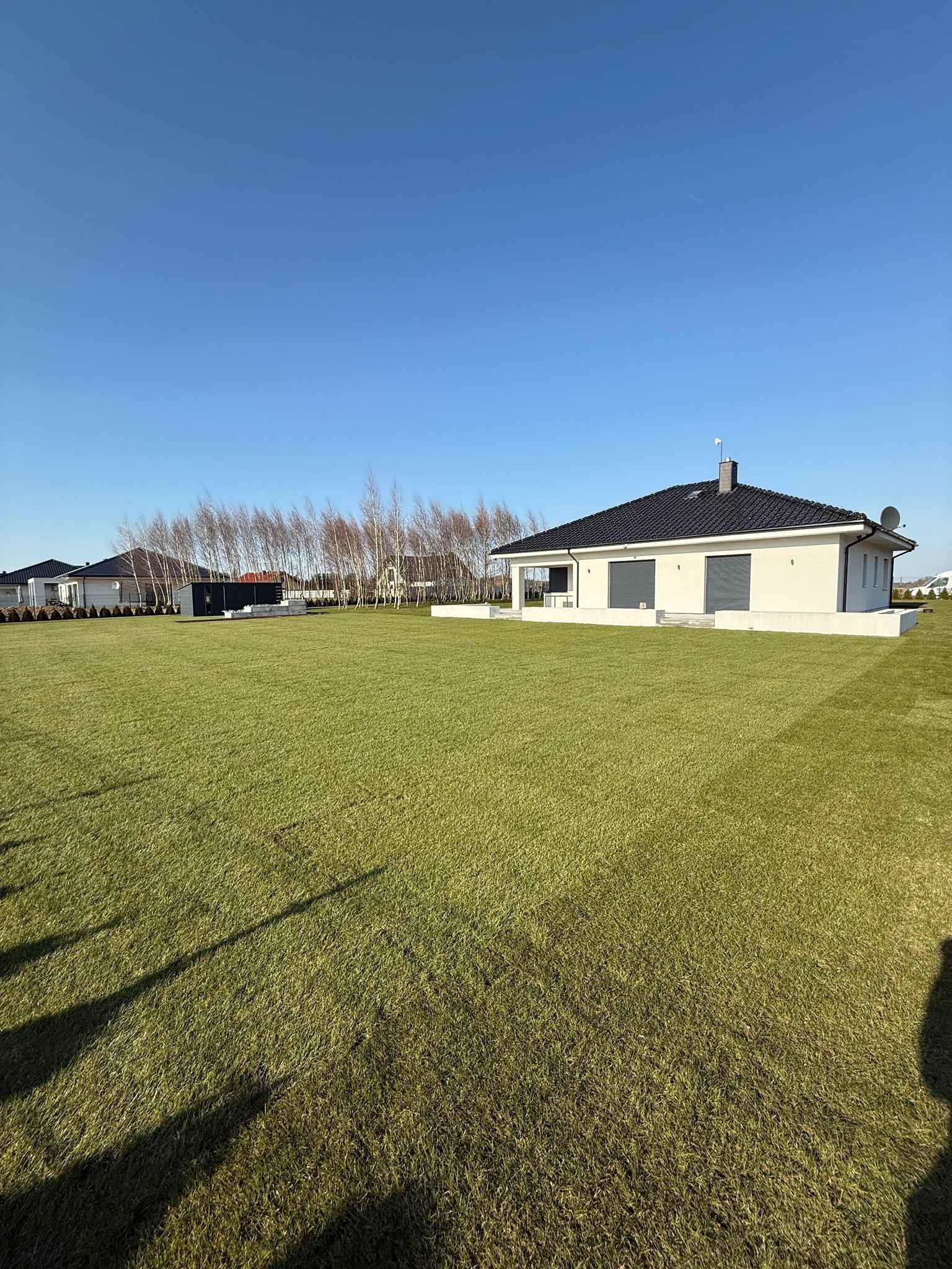 Rozległy trawnik przed nowoczesnym domem z roletami zewnętrznymi. W tle drzewa bez liści i błękitne niebo. Świeżo skoszona trawa w promieniach słońca.