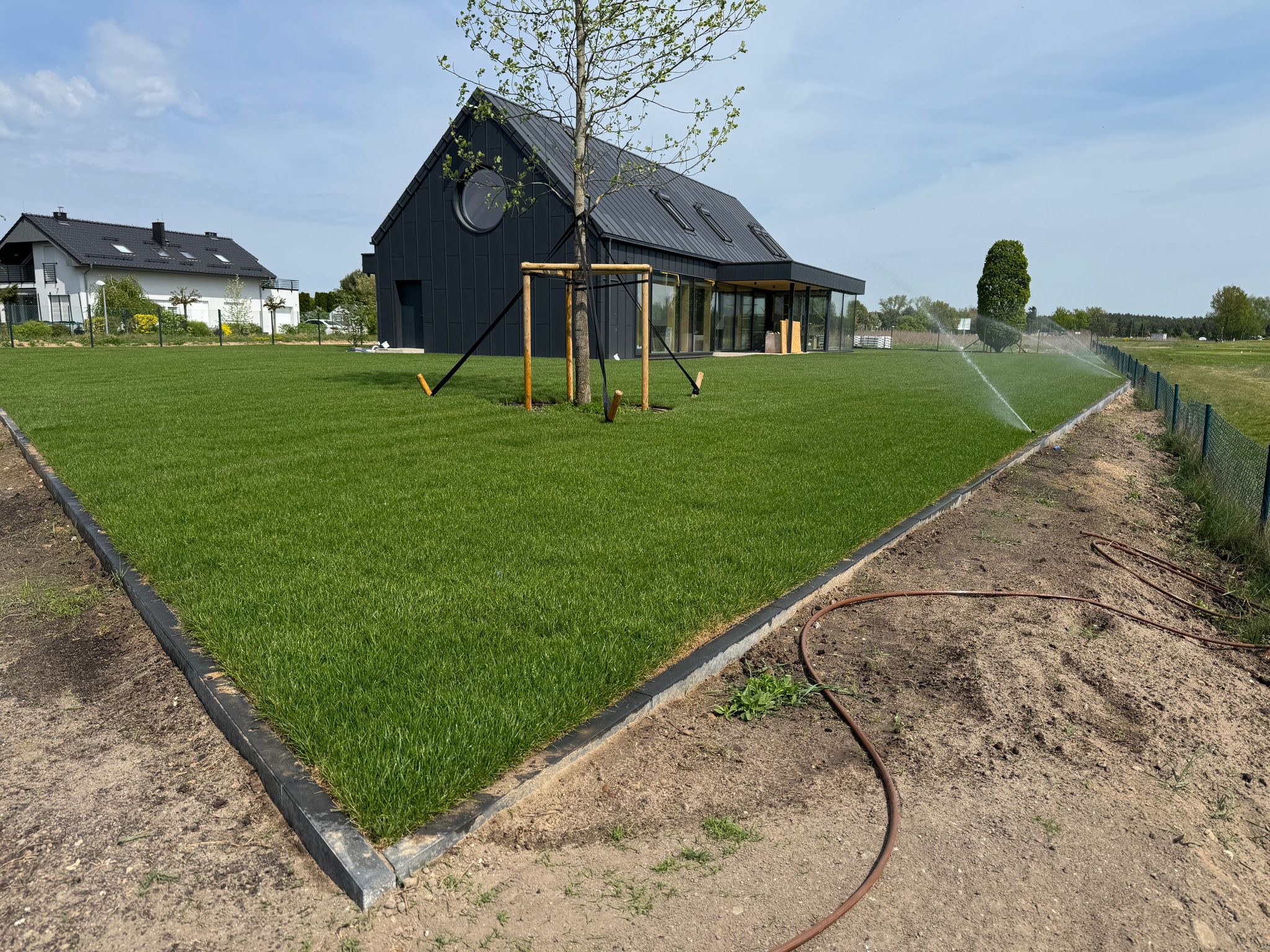 Nowoczesny dom z czarną elewacją i okrągłym oknem, otoczony świeżo założonym trawnikiem z rolki, nawadnianym przez zraszacze. Widoczne obrzeża trawnika i młode drzewko.