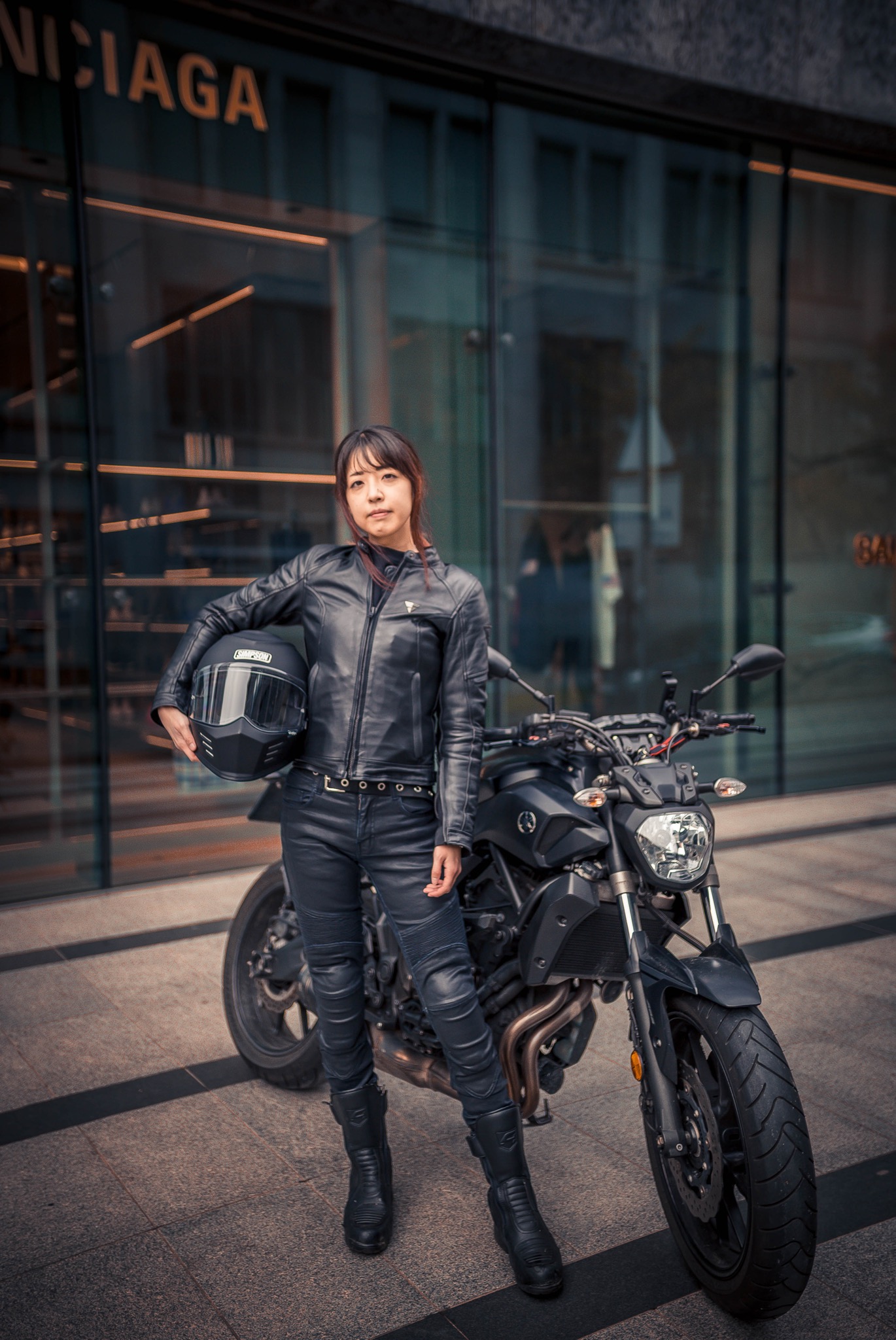 Kobieta w skórzanym stroju motocyklowym, trzymająca kask, pozuje przy czarnym motocyklu na tle szklanej witryny sklepu.