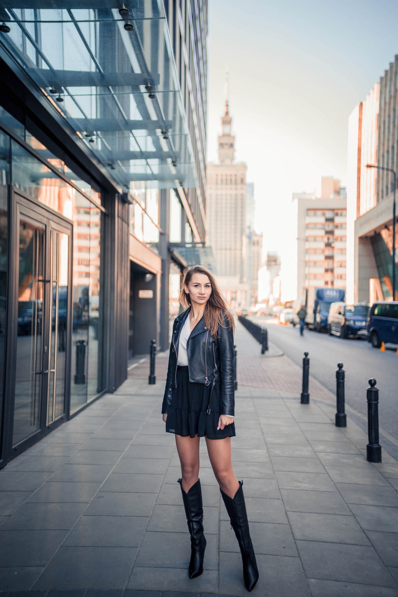 Kobieta w czarnej kurtce i spódnicy pozuje na tle nowoczesnego budynku z przeszkloną fasadą i Pałacu Kultury w oddali.