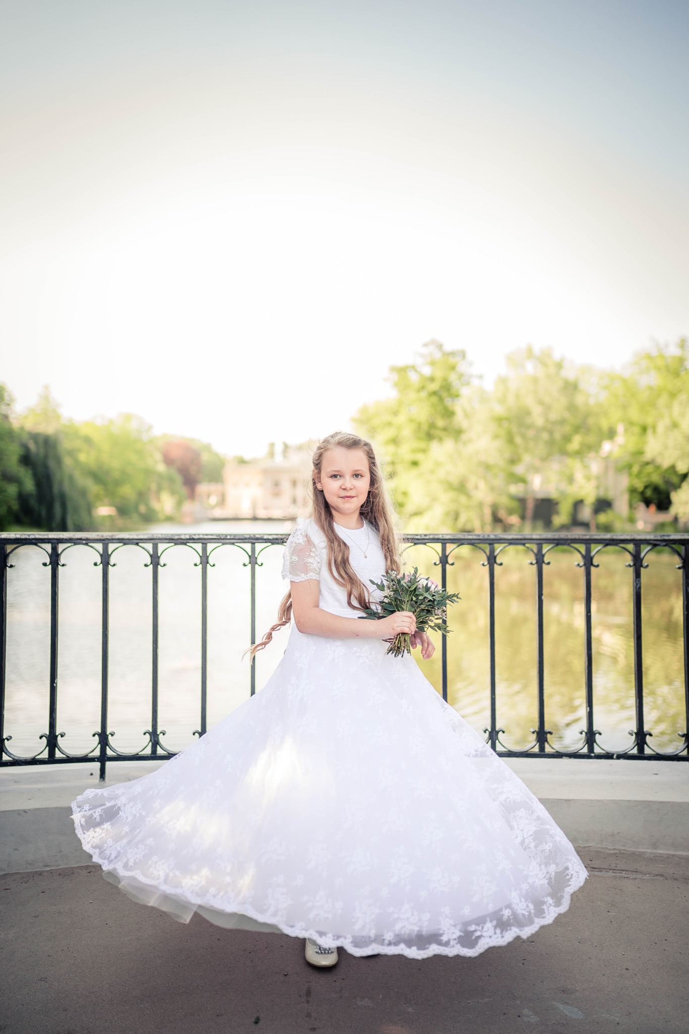 Dziewczynka w białej, koronkowej sukience komunijnej trzyma bukiet kwiatów na tle mostu i parku.