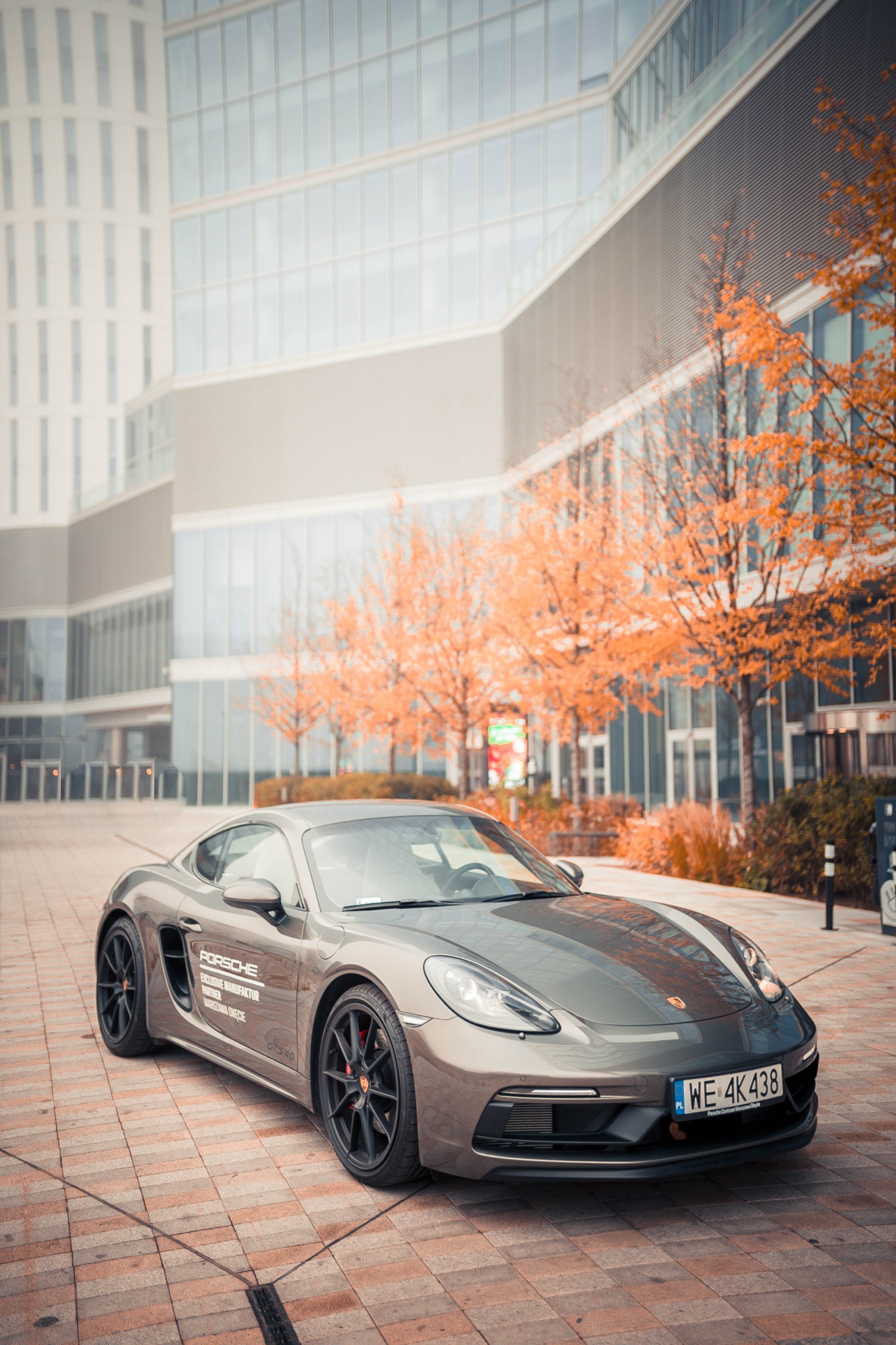 Szary sportowy samochód Porsche zaparkowany na brukowanym placu przed nowoczesnym budynkiem biurowym z pomarańczowymi drzewami w tle.