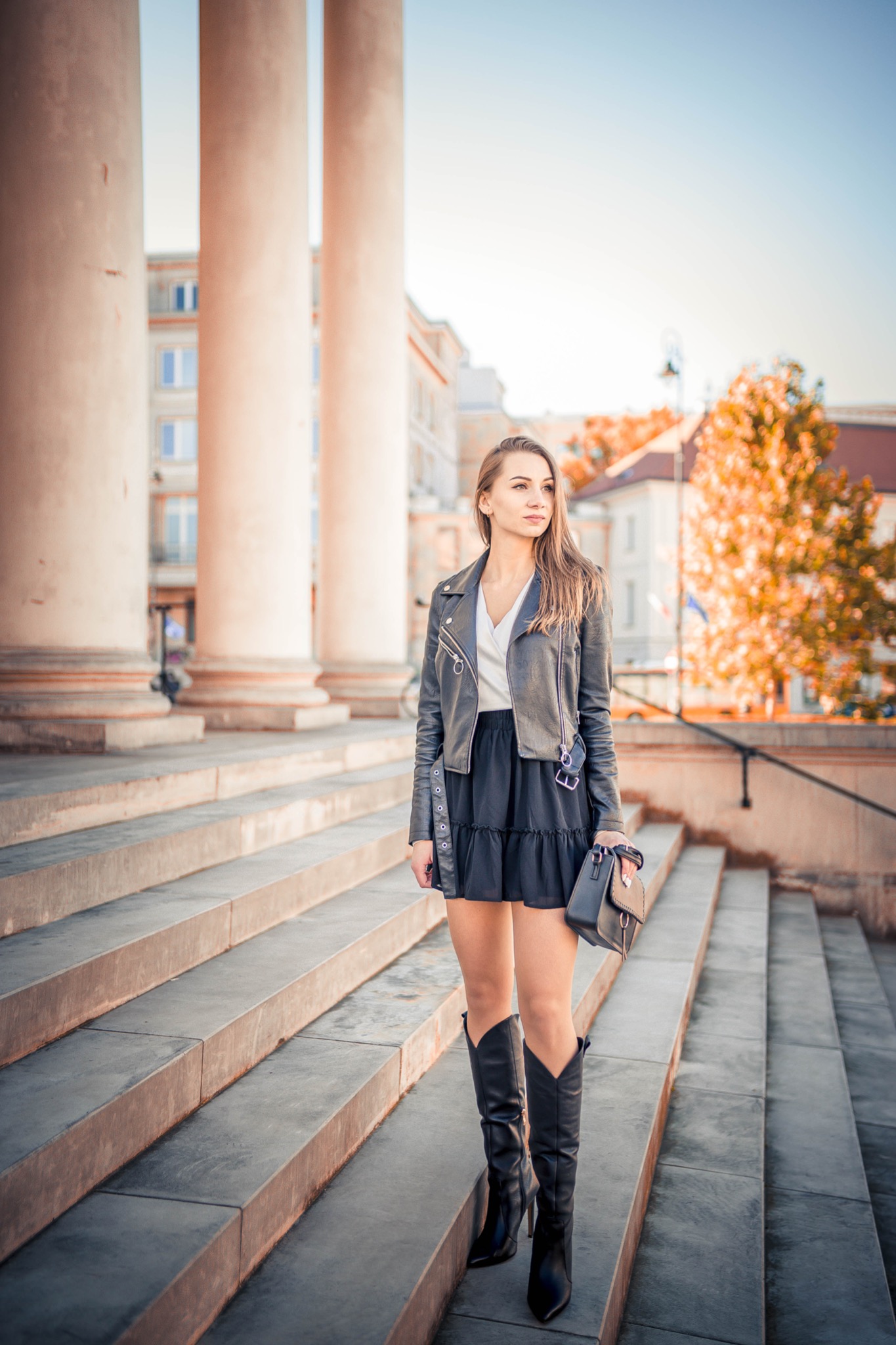 Kobieta w czarnej skórzanej kurtce i spódnicy pozuje na szerokich schodach z kolumnami w tle, trzymając małą torebkę.