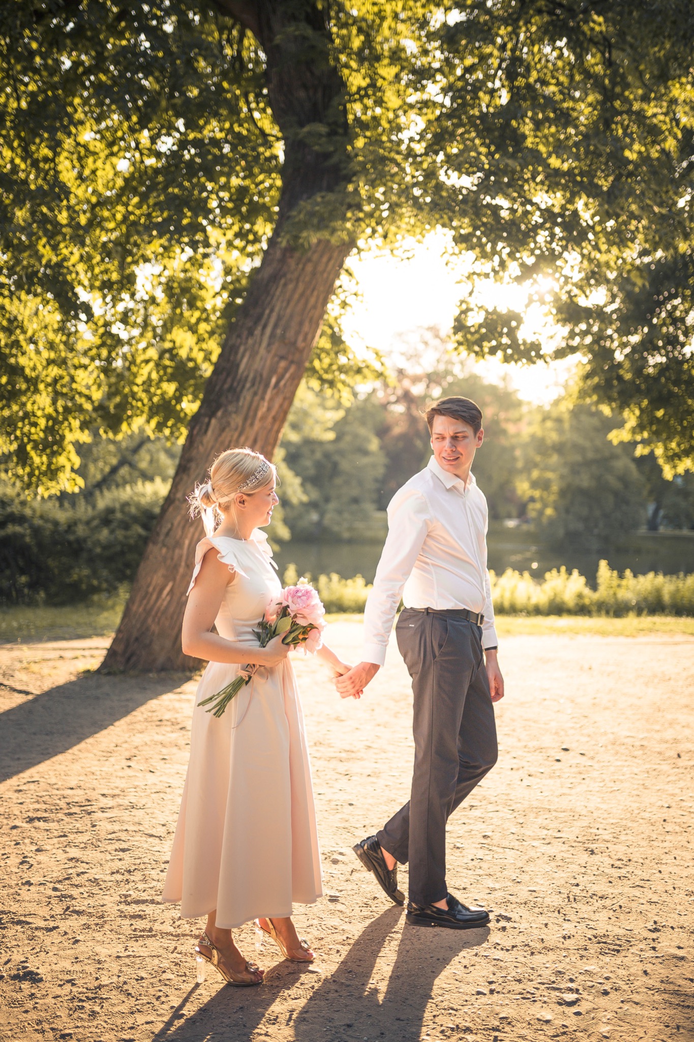 Para trzymająca się za ręce spaceruje w parku w promieniach zachodzącego słońca, kobieta w jasnej sukni z bukietem różowych piwonii, mężczyzna w białej koszuli i ciemnych spodniach.