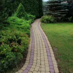 Kręta ścieżka z kostki brukowej w dwóch kolorach prowadzi przez zadbany ogród z różnorodną roślinnością, w tle wysokie, gęste tuje i świerk.