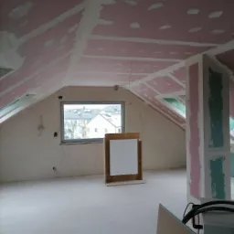 Poddasze w trakcie prac wykończeniowych: ściany i skośny sufit pokryte płytami z widocznymi łączeniami i szpachlowaniem, okno z widokiem na miasto, tymczasowa tablica informacyjna na drewnianym...