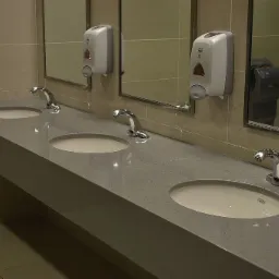 Długa, szara umywalka z trzema owalnymi zlewami, chromowanymi kranami, dozownikami mydła i lustrami w publicznej toalecie, widoczna suszarka do rąk w odbiciu.