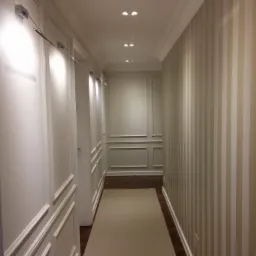 Korytarz z panelami ściennymi w kolorze białym i tapetą w beżowo-białe pasy, oświetlony punktowo i kinkietami, z widoczną podłogą z ciemnego drewna i beżowym dywanem.