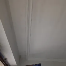 Wykończony sufit z listwami dekoracyjnymi, świeżo po malowaniu na biało, widoczne zabezpieczone okno folią malarską.