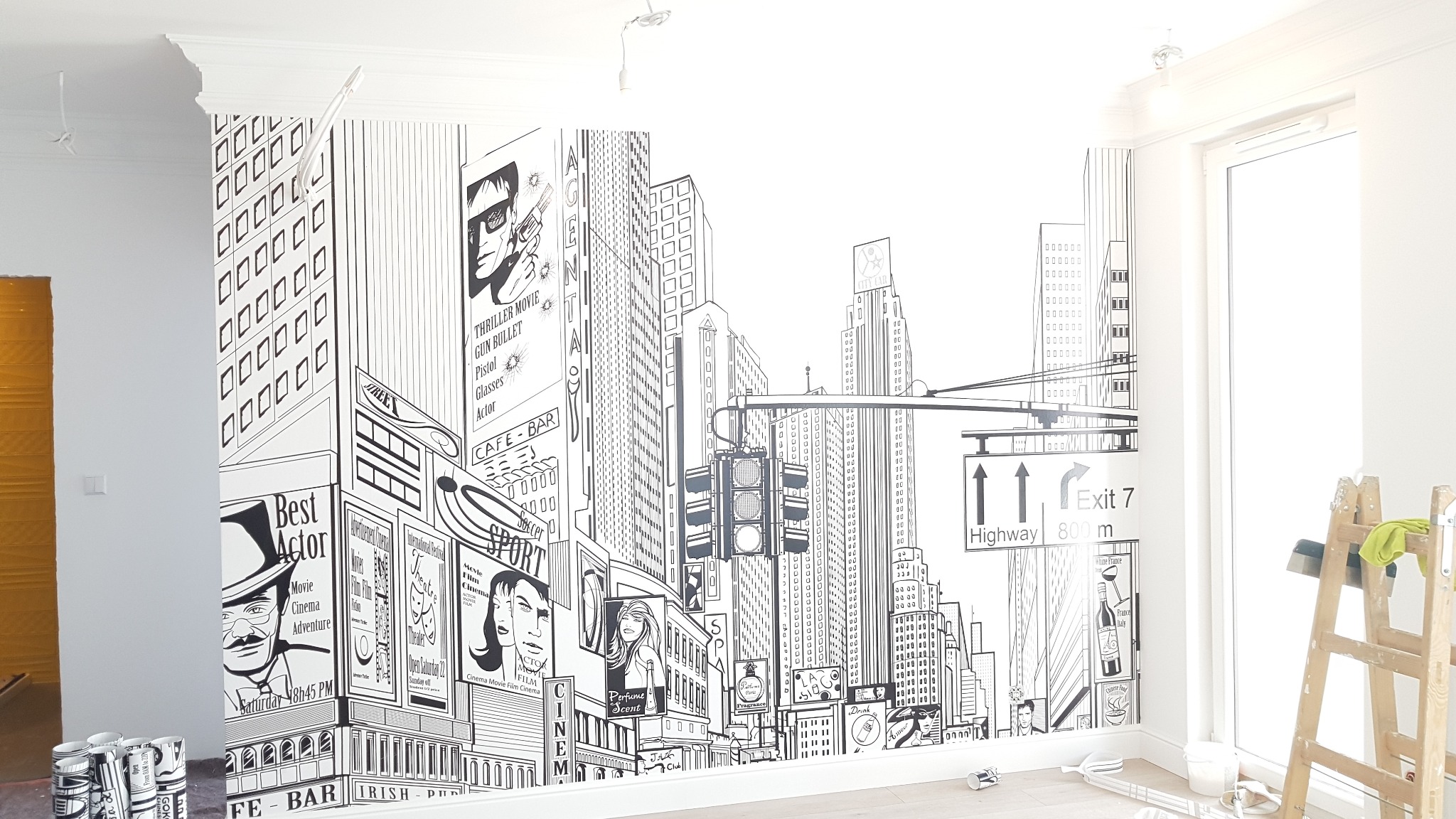 Ściana w pokoju po tapetowaniu z motywem czarno-białej panoramy miasta, widoczne puszki po kleju i drabina malarska.