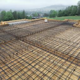 Firma budowlana DZIADOŃ - Zbrojenie płyty fundamentowej z drewnianym szalunkiem na tle mglistego krajobrazu z zabudowaniami i zielenią.