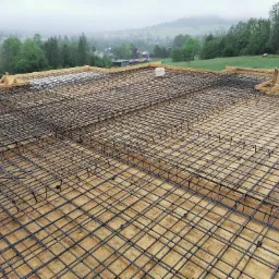 Zbrojenie płyty fundamentowej z drewnianym szalunkiem na tle mglistego krajobrazu z zabudowaniami i zielenią.