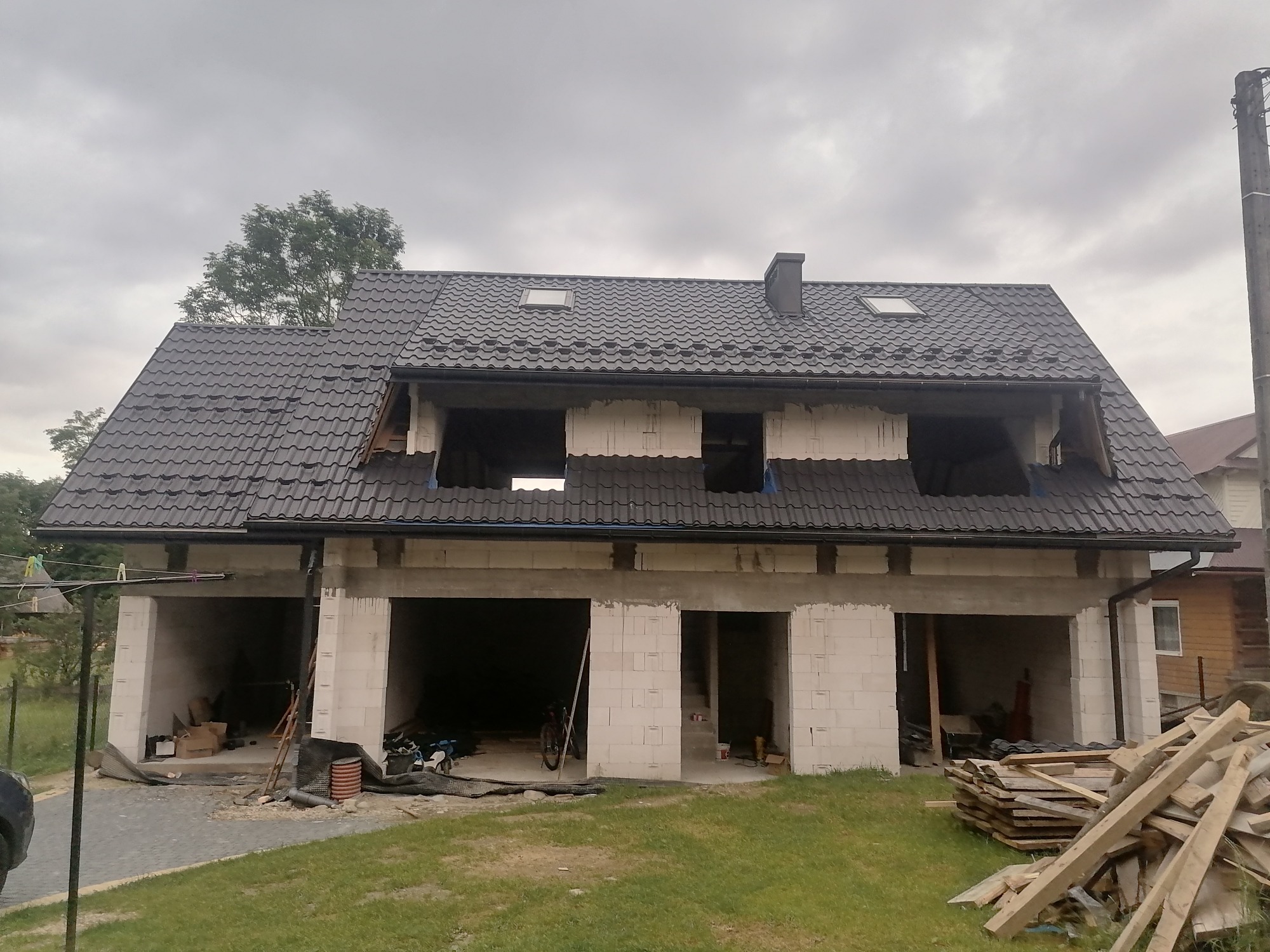 Dwukondygnacyjny budynek w stanie surowym otwartym z ciemną dachówką, widoczne otwory okienne i drzwiowe, na pierwszym planie stos desek, pochmurne niebo.