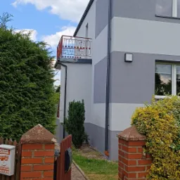 Dwukondygnacyjny dom z balkonem, pomalowany w poziome pasy szarości, widoczny zza ceglanego ogrodzenia z otwartą furtką i skrzynką na listy.