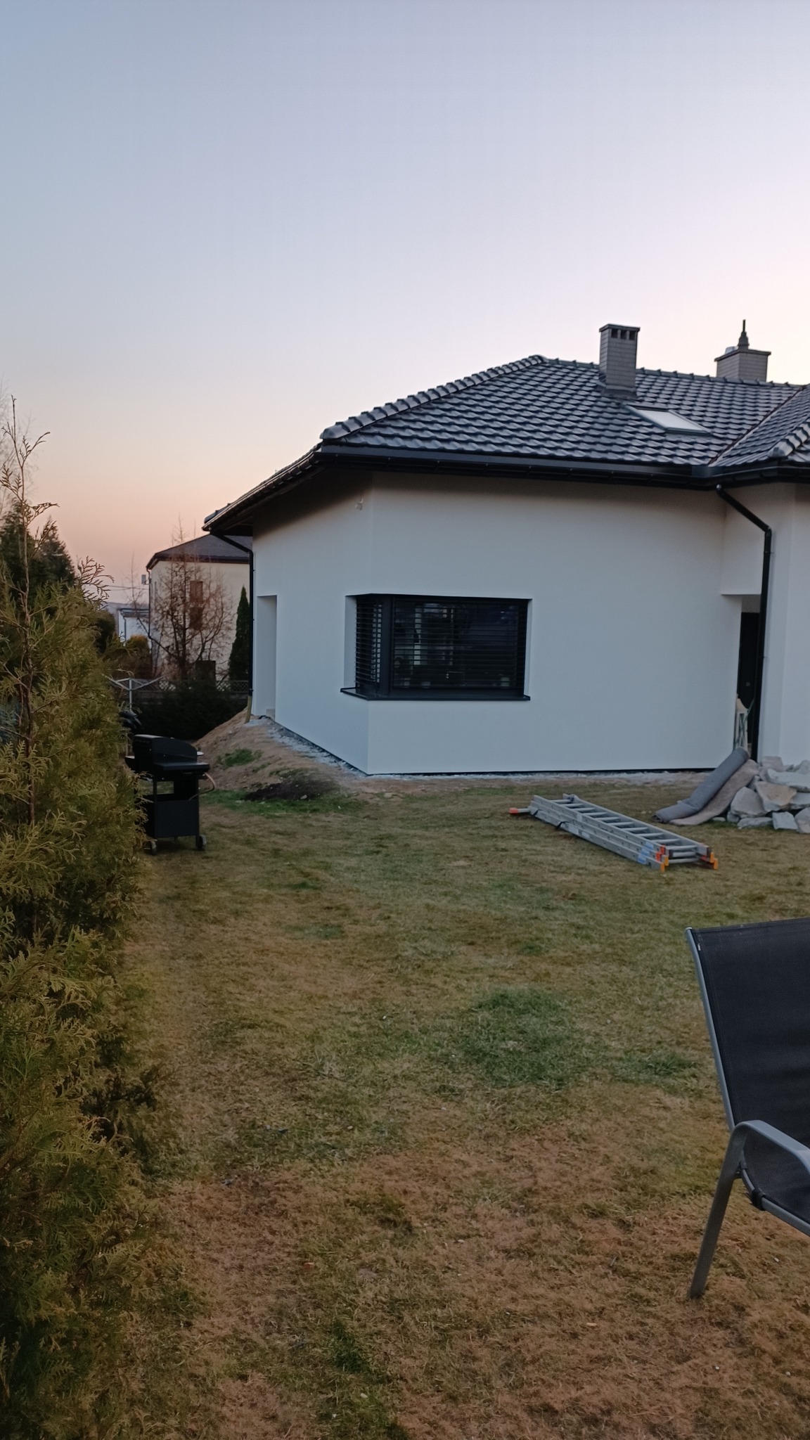 Nowoczesny dom z białą elewacją i ciemną dachówką, widok z ogrodu z grillem, drabina oparta o ścianę, świeżo obsypana ziemia przy fundamencie.