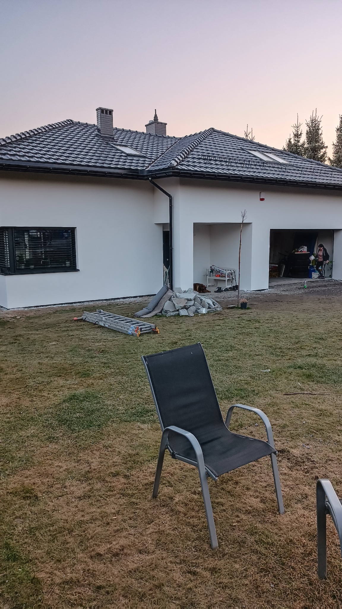 Nowoczesny dom z białą elewacją i szarą dachówką, widoczny z perspektywy ogrodu, z rozłożoną drabiną i stertą kamieni obok ściany, sugerujące prace wykończeniowe elewacji.