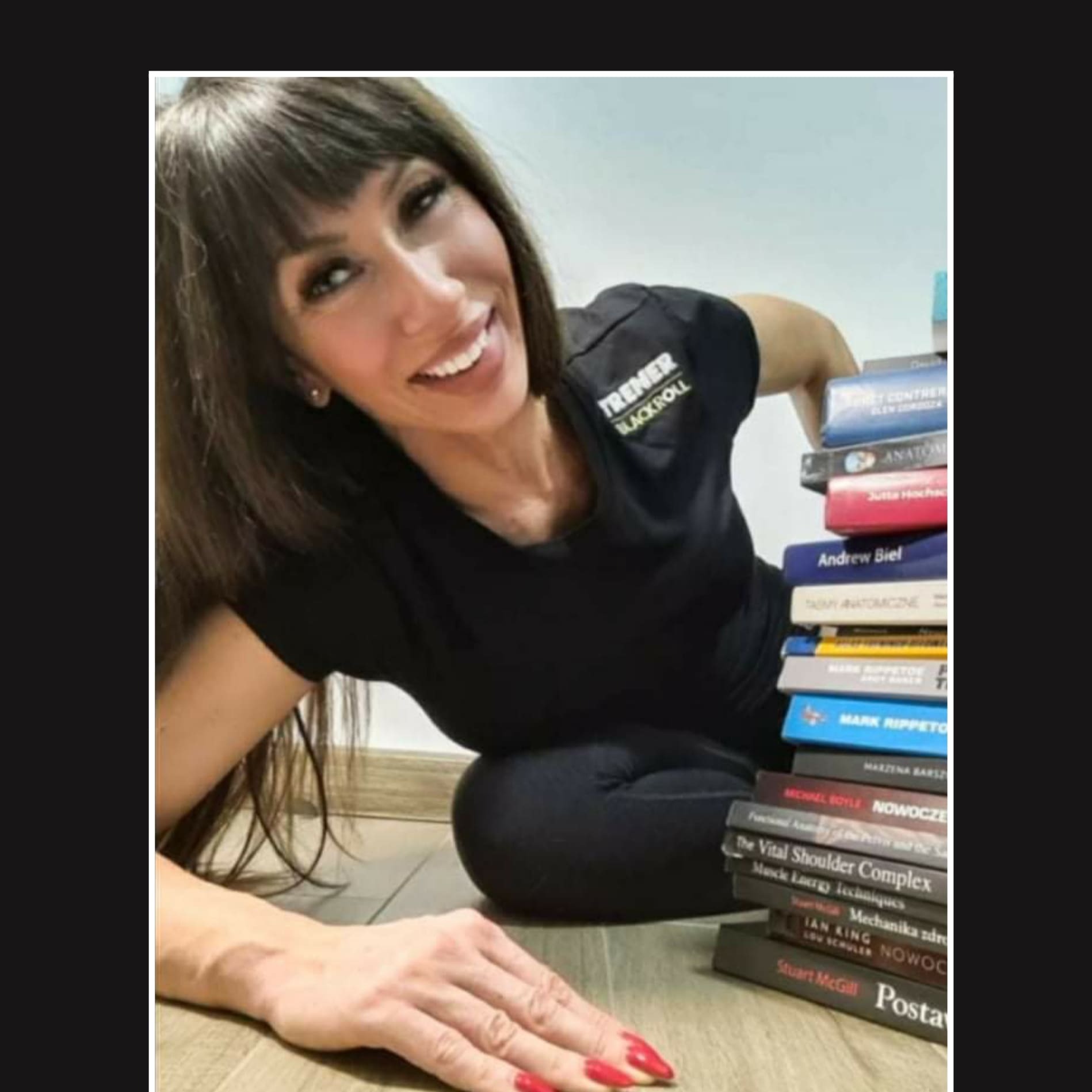 Uśmiechnięta kobieta w czarnej koszulce z napisem 'TRENER BLACKROLL' pozuje obok stosu książek o anatomii i treningu, leżąc na podłodze.