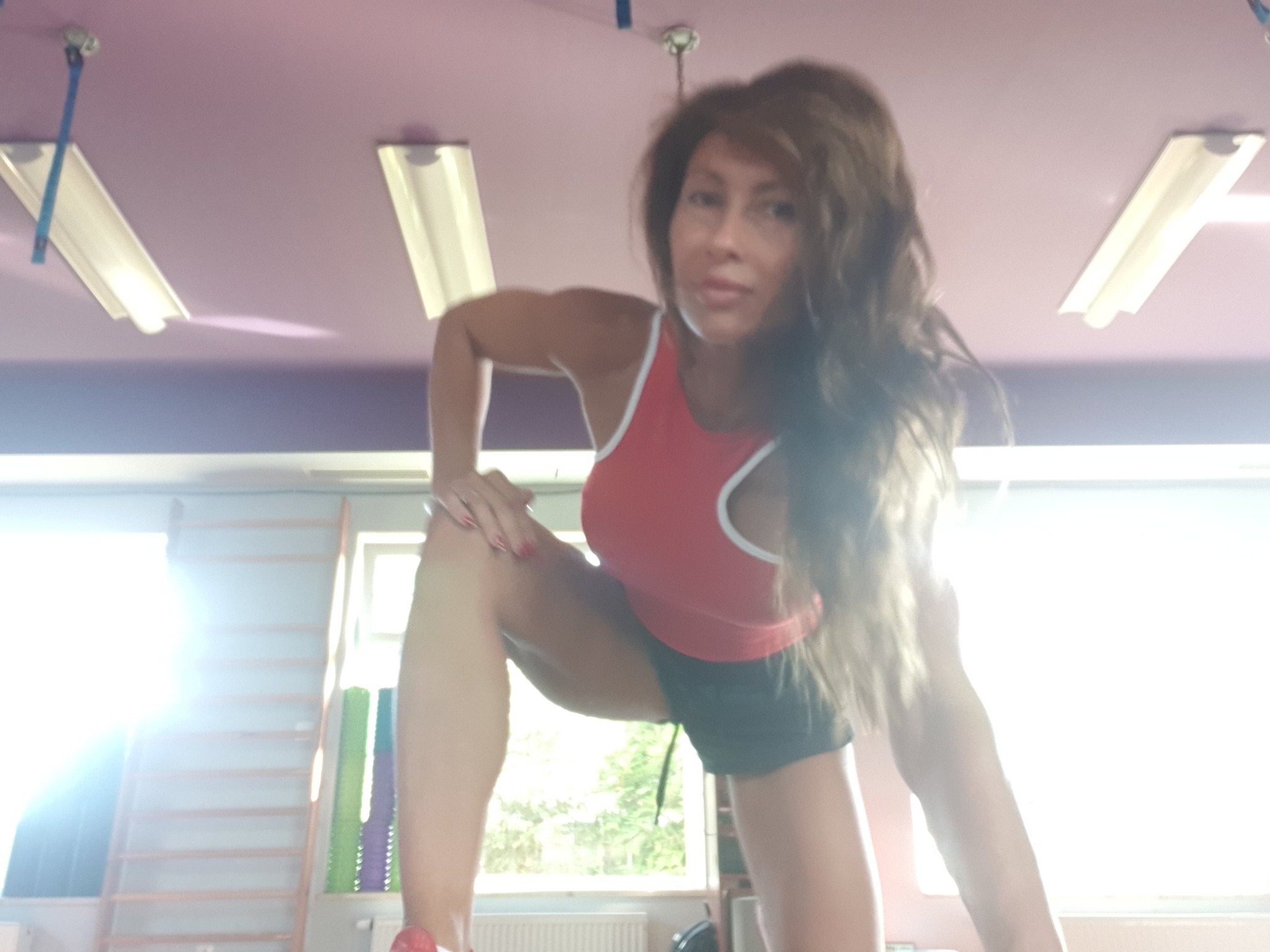 Kobieta w sportowym stroju wykonuje rozciąganie w jasnym pomieszczeniu z drabinkami gimnastycznymi i fioletowym sufitem.