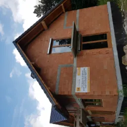 Budynek w trakcie budowy z zamontowanymi oknami w brązowych ramach, widoczny baner reklamowy firmy oferującej okna, drzwi i bramy garażowe.