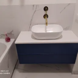 Nowoczesna łazienka z białą umywalką nablatową, złotą armaturą podtynkową i szafką w kolorze granatowym, marmurowe płytki na ścianie.