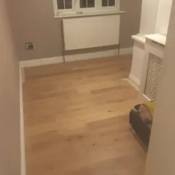 Jasny, drewniany parkiet w pokoju po odświeżeniu, ściany pomalowane na szaro, widoczne gniazdko elektryczne bez osprzętu, biały grzejnik pod oknem.