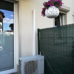 Zewnętrzna jednostka klimatyzacji zamontowana na drewnianym tarasie obok okna z żaluzjami, z wiszącą donicą z fioletowymi kwiatami w tle.