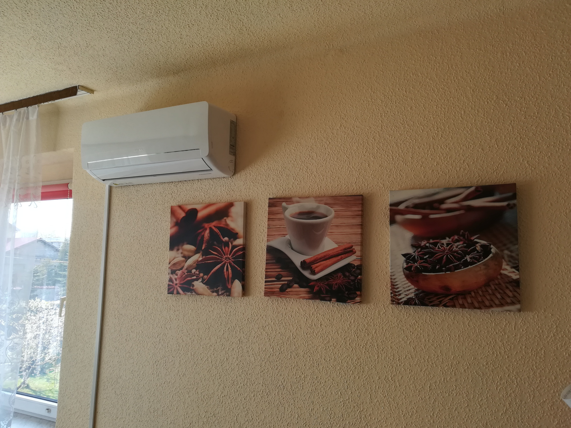 Biały klimatyzator ścienny zamontowany w pokoju z beżową, teksturowaną ścianą, obok trzy obrazy z motywami kawy i przypraw.