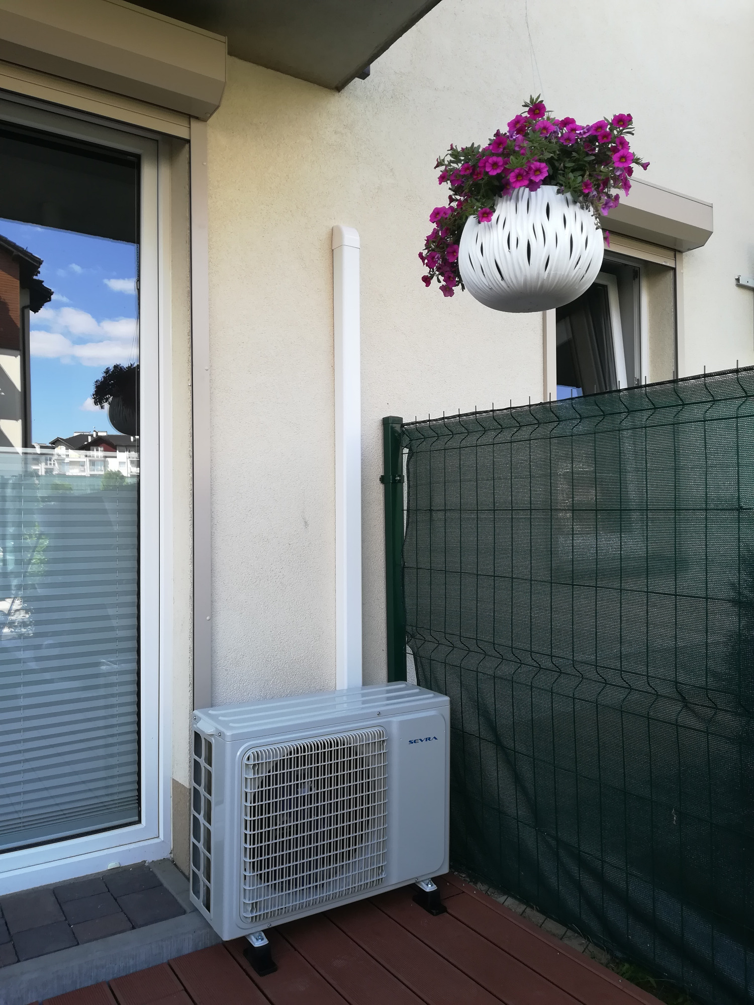 Zewnętrzna jednostka klimatyzacji zamontowana na drewnianym tarasie obok okna z żaluzjami, z wiszącą donicą z fioletowymi kwiatami w tle.
