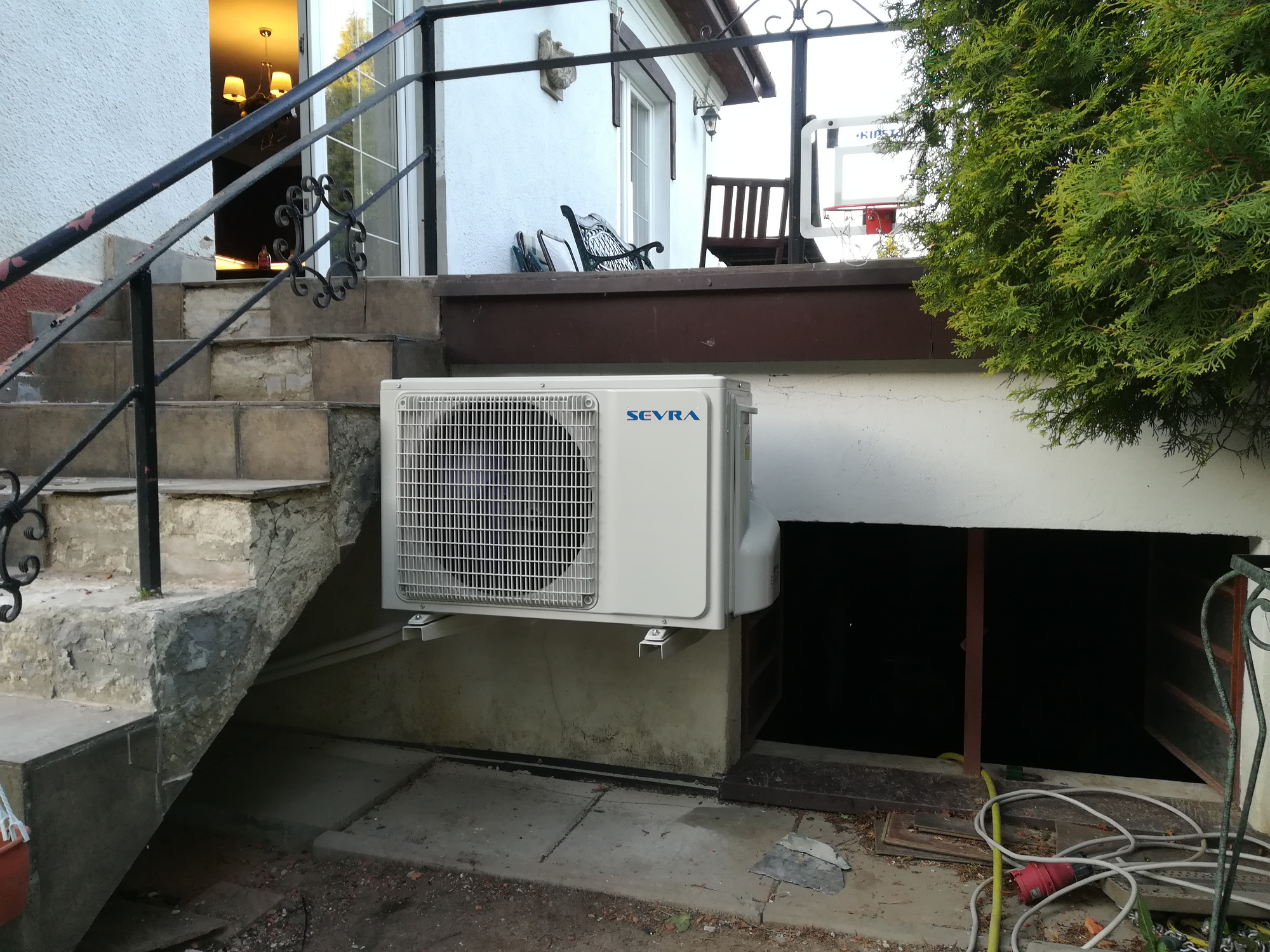 Zewnętrzna jednostka klimatyzacji marki Sevra zamontowana na ścianie budynku, widoczna z perspektywy schodów prowadzących do wejścia.