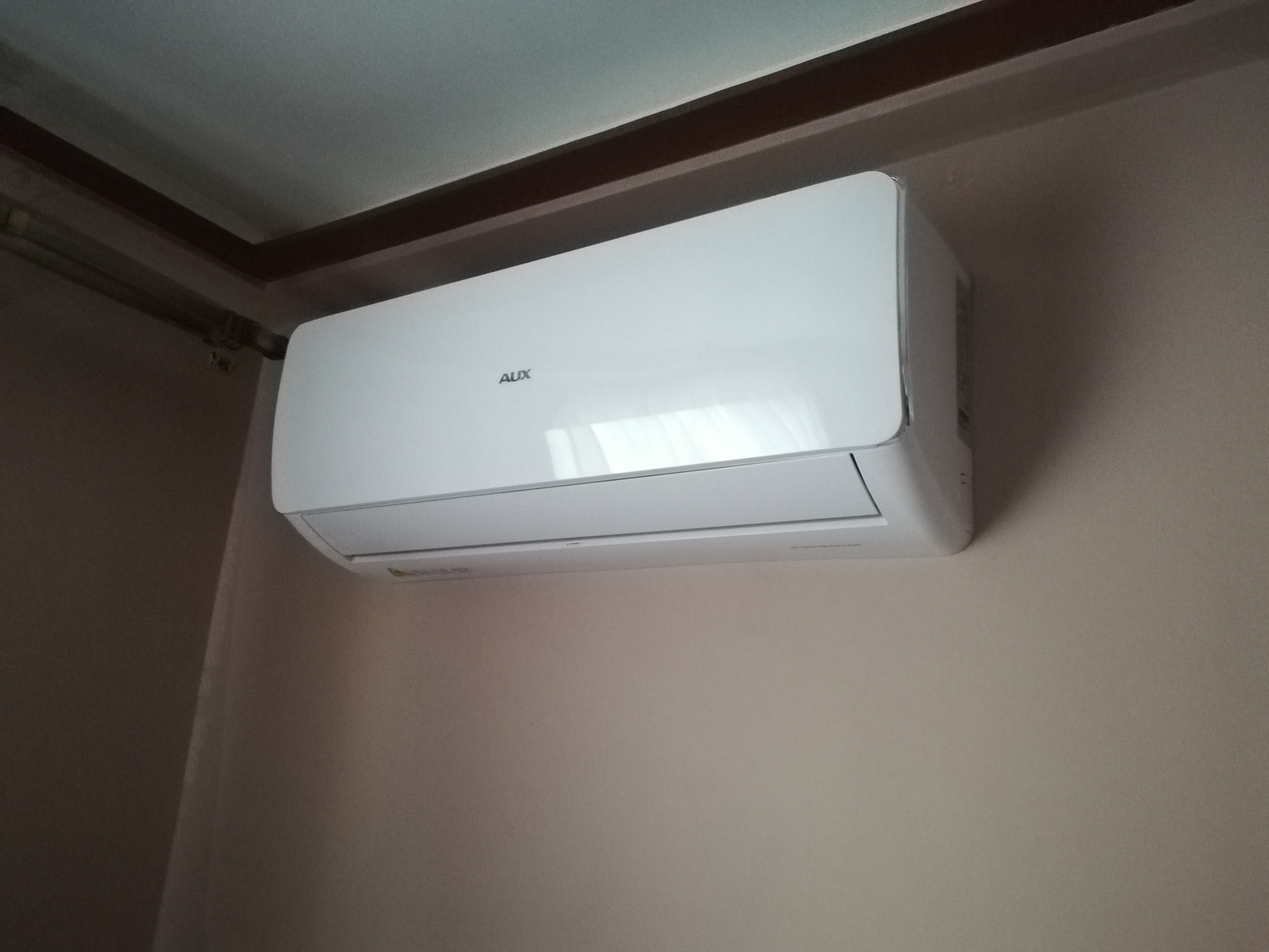 Biała jednostka klimatyzacji AUX zamontowana na ścianie w pomieszczeniu z beżowymi ścianami i brązowymi listwami sufitowymi.