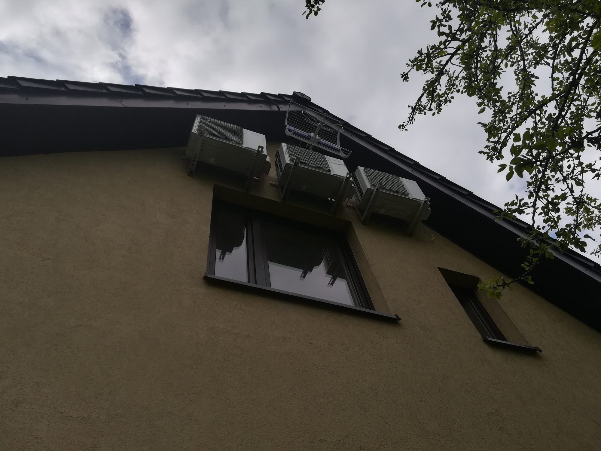 Zewnętrzne jednostki klimatyzacji zamontowane na elewacji domu jednorodzinnego, widok z dołu, szara elewacja, ciemne okna, fragment dachu i drzewa na tle pochmurnego nieba.