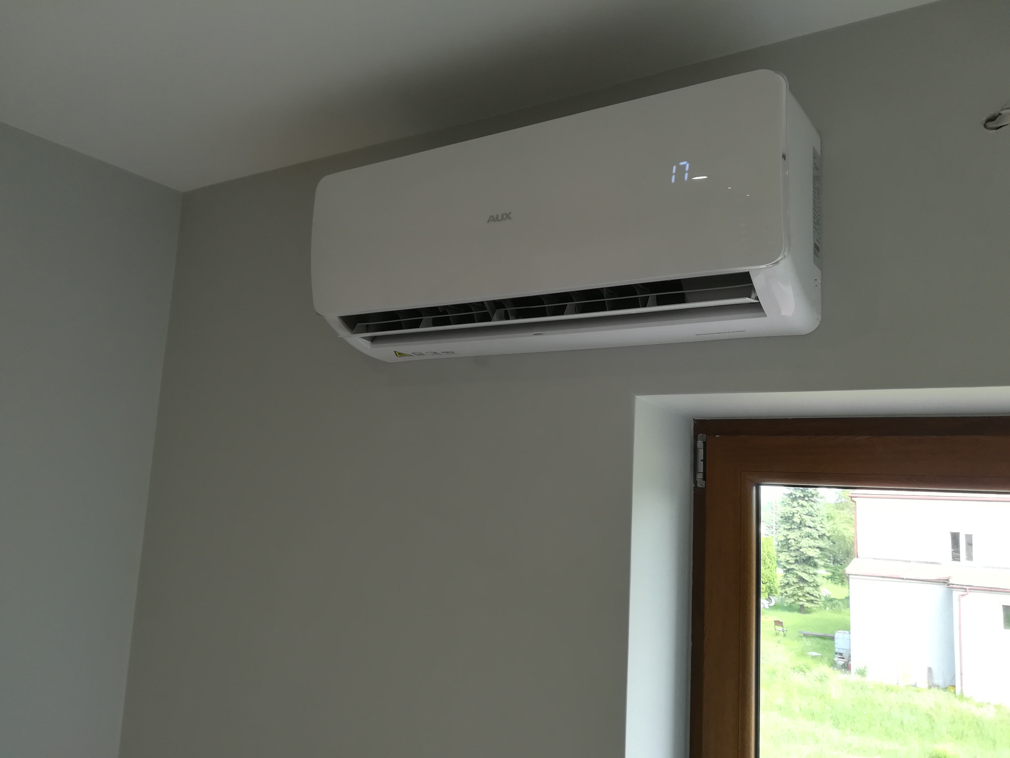 Biały klimatyzator marki AUX zamontowany na szarej ścianie w pomieszczeniu, widok z okna na zieleń i budynek.
