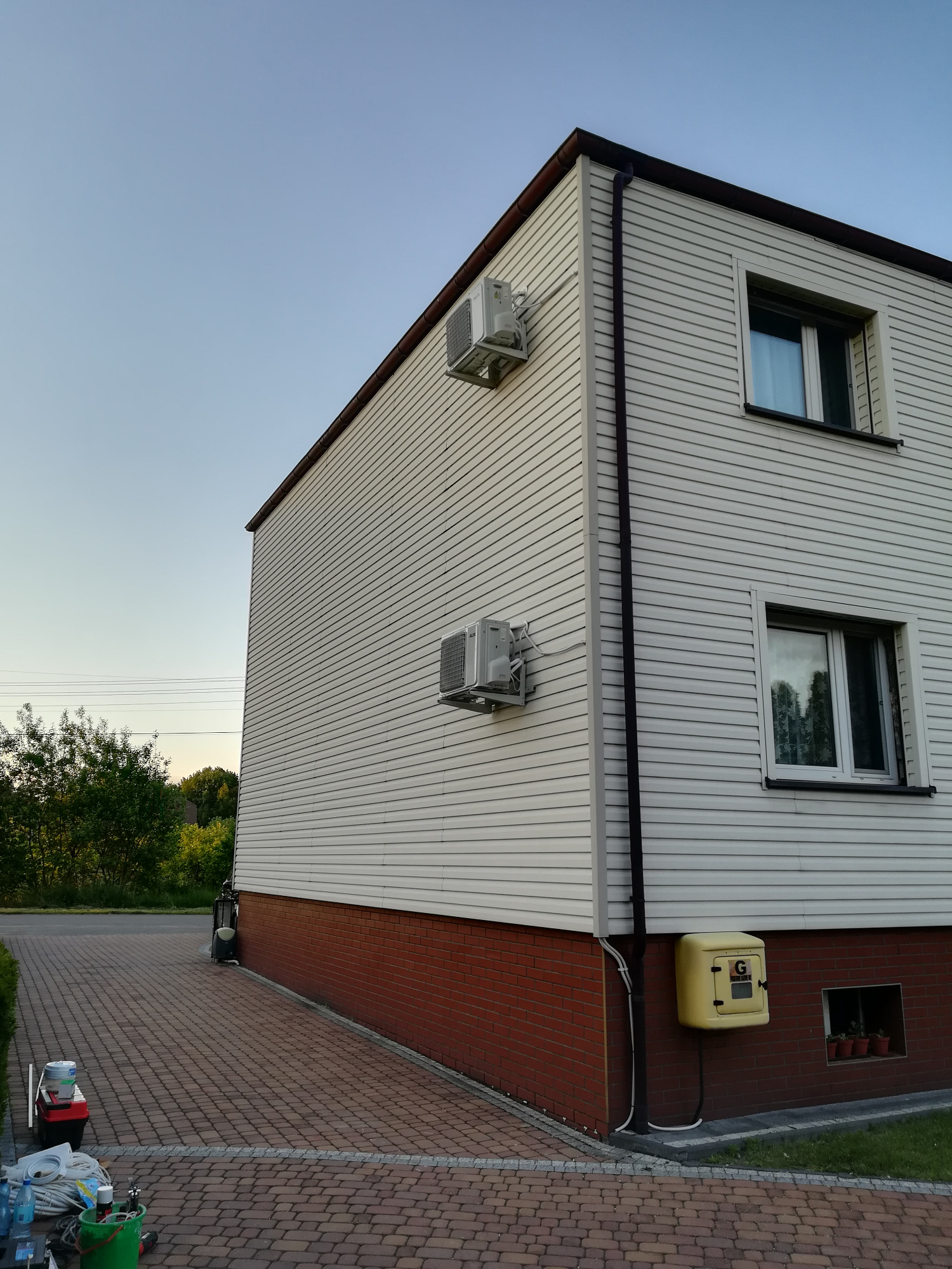 Dwa klimatyzatory zamontowane na bocznej ścianie domu z sidingiem, widok z poziomu brukowanego podjazdu, z widocznymi narzędziami i materiałami budowlanymi sugerującymi niedawny montaż.