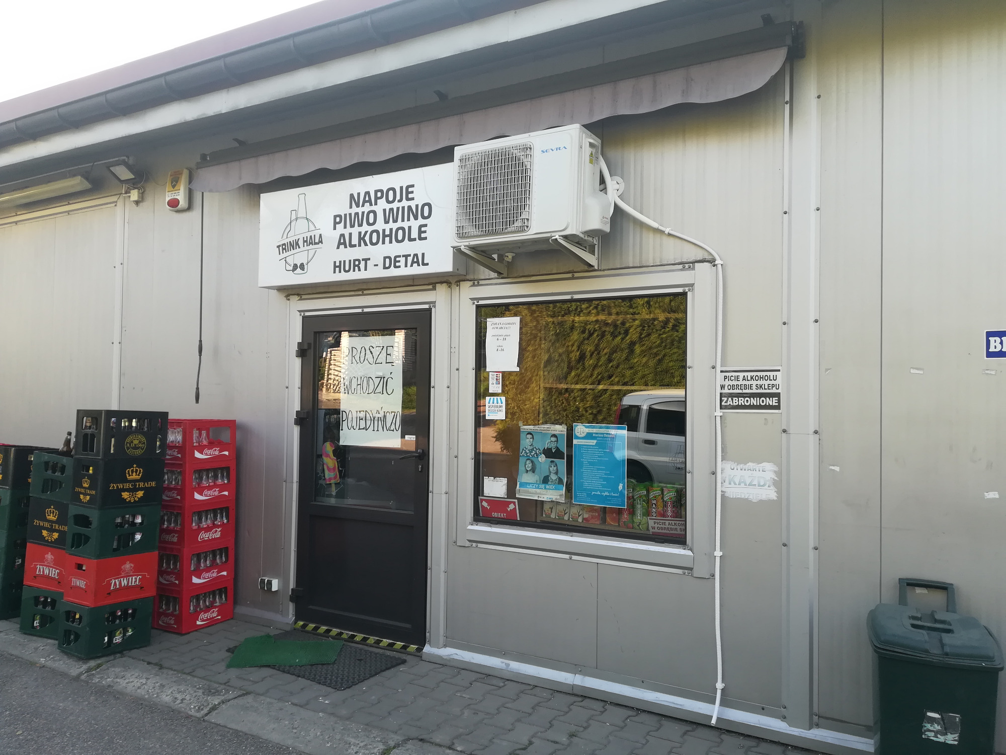 Zewnętrzny klimatyzator zamontowany nad witryną sklepu monopolowego, widoczne skrzynki z napojami i szyld z napisem 'Napoje, piwo, wino, alkohole'.
