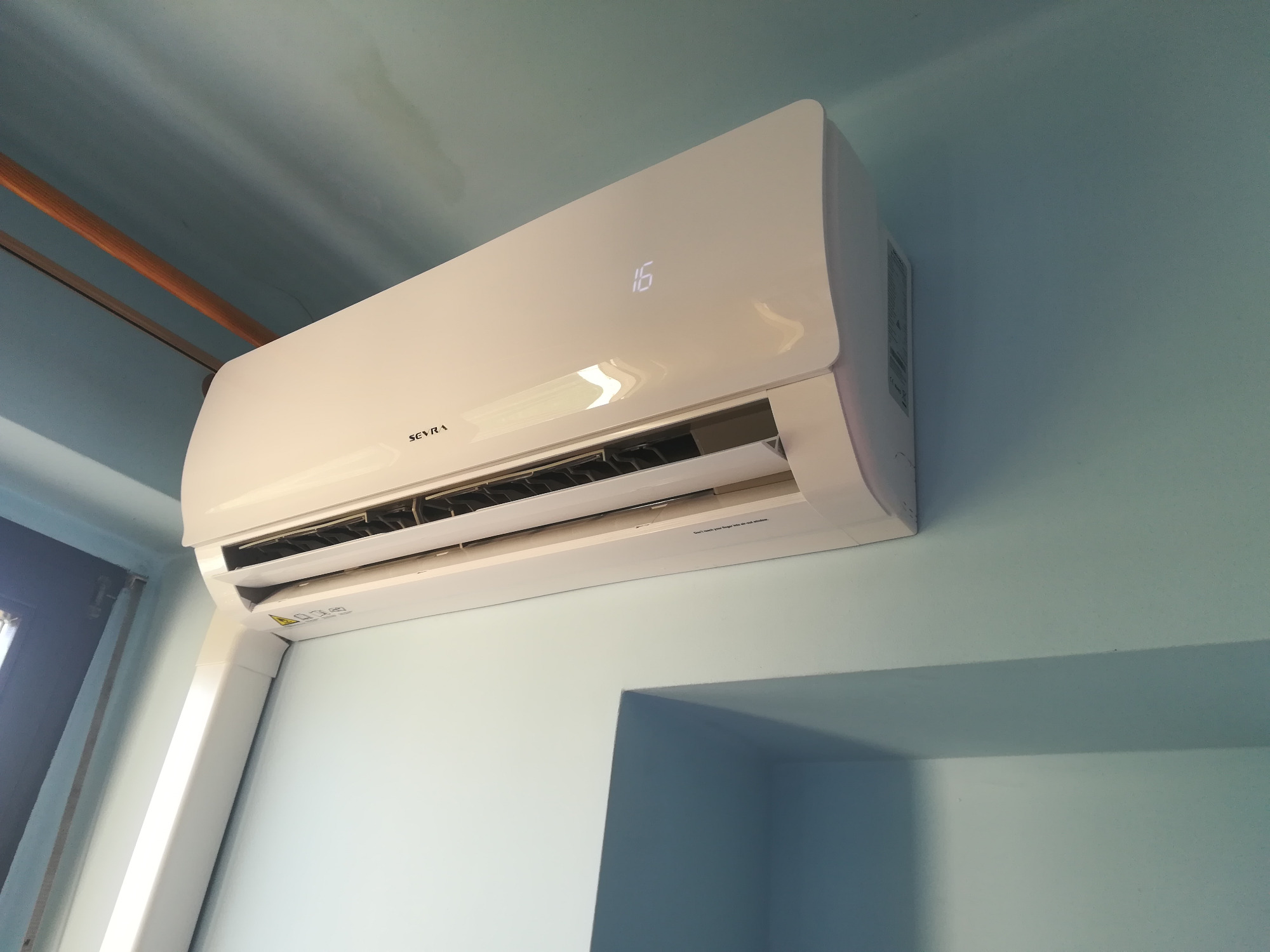 Biały klimatyzator marki Sevra zamontowany na błękitnej ścianie, wyświetlający temperaturę 16 stopni Celsjusza.